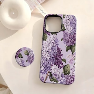 May include: A phone case and matching phone grip featuring a lilac floral design in shades of purple and white. The case has a black camera cutout. The grip is round and has the same floral pattern. The background is a light purple.