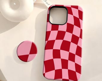 Puede incluir: Funda de teléfono y agarre a juego a cuadros rosas y rojos. La funda tiene un recorte negro para la cámara y un patrón de tablero de ajedrez ondulado. El agarre del teléfono es redondo y bicolor.