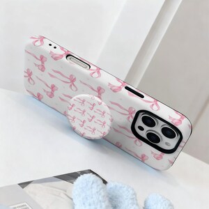 May include: White phone case with a pink bow pattern and a matching pop socket. The case has a black camera module and a white background. The pop socket is round and has a repeating pattern of pink bows.