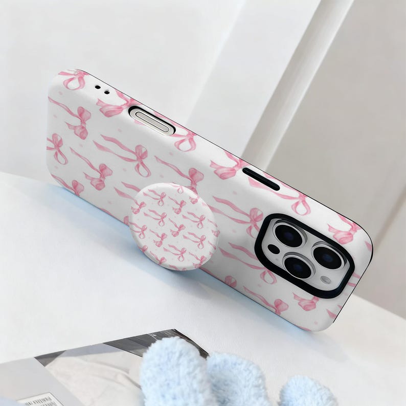 May include: White phone case with a pink bow pattern and a matching pop socket. The case has a black camera module and a white background. The pop socket is round and has a repeating pattern of pink bows.