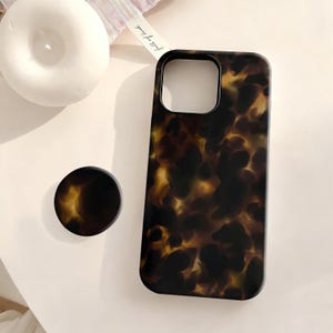 May include: A phone case and matching phone grip with a tortoiseshell pattern in shades of brown and amber. The phone case has a camera cutout and a smooth, glossy finish. The grip is round and flat.