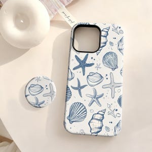May include: White phone case and pop socket with a blue seashell and starfish pattern. The phone case has a camera cutout and a smooth finish. The pop socket is round and has a matching design. A white candle is in the background.