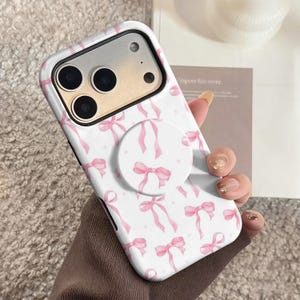 May include: White phone case with a pink bow pattern and a matching pop socket. The case has a glossy finish and a protective bumper. The pop socket is round and features a pink bow design. The phone case is held in a person's hand.