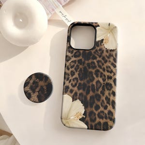 Puede incluir: Una funda de teléfono y un pop socket a juego con un diseño de estampado de leopardo y acentos florales color crema. La funda del teléfono es negra alrededor de la cámara y tiene un acabado brillante. El pop socket es redondo y tiene el mismo estampado de leopardo.