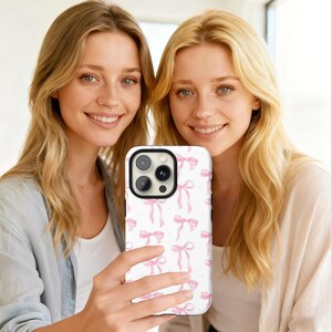 May include: A white phone case featuring a pattern of pink bows and small dots. The case has a black camera module and is held by a person. The background shows two people with blonde hair.