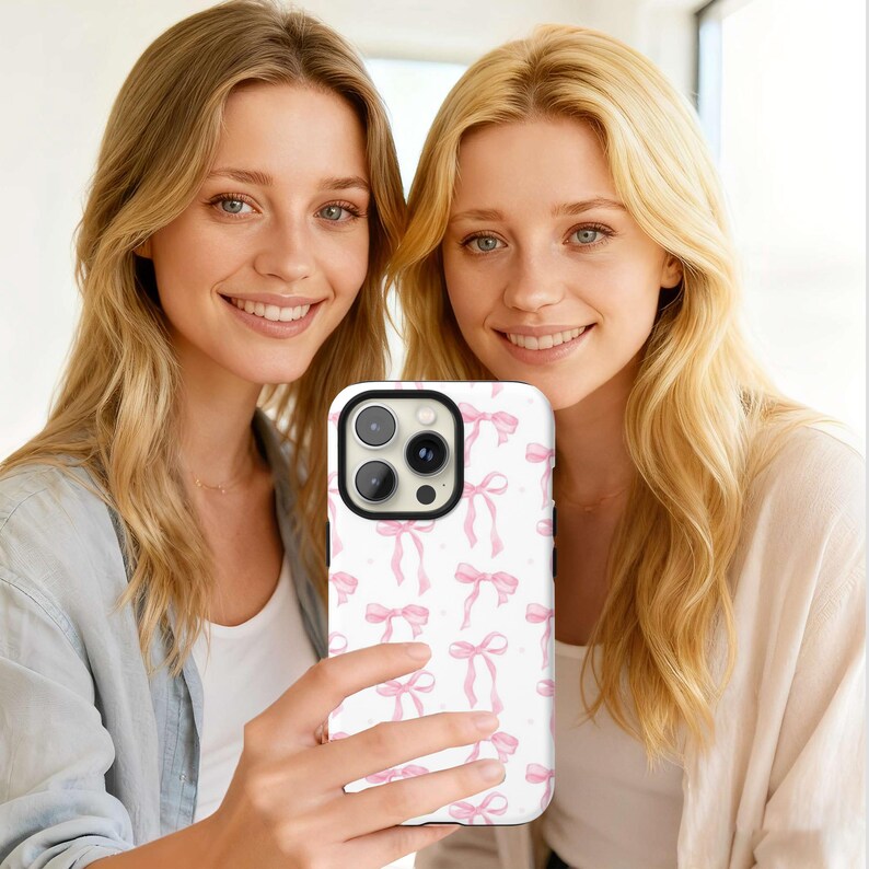 May include: A white phone case featuring a pattern of pink bows and small dots. The case has a black camera module and is held by a person. The background shows two people with blonde hair.