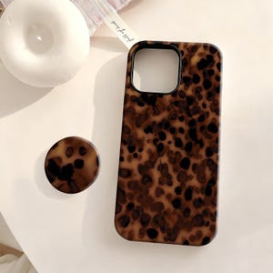 May include: A brown and black patterned phone case and matching phone grip. The case has a rectangular shape with a camera cutout. The grip is round. The pattern resembles a tortoiseshell design.