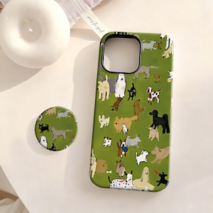 Può includere: Una custodia per telefono verde e un supporto per telefono abbinato con un motivo ripetuto di varie razze di cani in diversi colori. La custodia ha un bordo nero attorno al ritaglio della fotocamera. L'impugnatura del telefono è rotonda e presenta lo stesso motivo di cani.