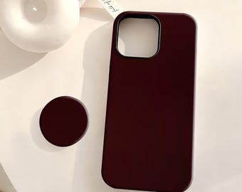 May include: A burgundy phone case and matching phone grip. The phone case has a square camera cutout and a smooth finish. The phone grip is a small, round disc. The items are on a white surface.
