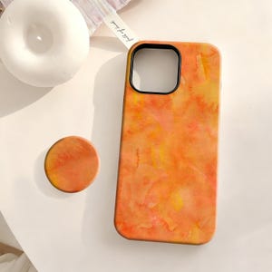 May include: Orange and yellow watercolor-style phone case with a matching round phone grip. The case has a black camera cutout. The phone grip is a circular shape. The background is a white surface.