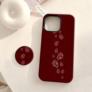 May include: A burgundy phone case and matching pop socket featuring a pattern of red ladybugs with black spots. The case has a camera cutout and a smooth finish. The pop socket is round and has the same ladybug design.