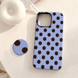 Pode incluir: Uma capa de telefone azul claro e um suporte de telefone correspondente com um padrão repetido de bolinhas marrons escuras. A capa do telefone tem um recorte de câmera preto. O suporte do telefone é redondo e tem duas bolinhas marrons escuras.