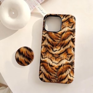 May include: A phone case and matching phone grip featuring a tiger stripe pattern in shades of brown and black. The phone case has a black border around the camera and screen. The phone grip is round and has the same tiger stripe design.