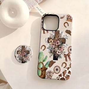 May include: A phone case and matching circular grip featuring a beige background with a pattern of wooden crosses adorned with flowers, green cacti, brown cowboy boots, hats, horseshoes, wagon wheels, playing cards, and a cow print corner.