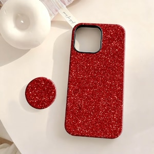 May include: A red glitter phone case and matching pop socket. The phone case has a black camera cutout and a shiny, textured surface. The pop socket is round and has a similar glittery finish. The items are on a white surface.