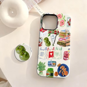 May include: A white phone case with a colorful illustration of groceries, books, and flowers. The design includes images of apples, strawberries, oranges, and shopping bags. A matching pop socket with green apples is also visible.