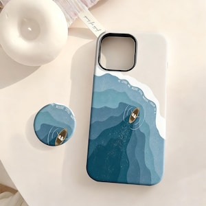 May include: A phone case and matching circular phone grip featuring a blue ocean wave design with a small boat and the text He leads me beside still waters Psalm 23:2. The case has a black camera cutout and a light beige upper section.