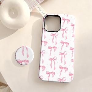 May include: White phone case and pop socket featuring a pattern of pink watercolour bows and small dots. The phone case has a black camera cutout. The pop socket has a matching design. A white candle and a ribbon are in the background.