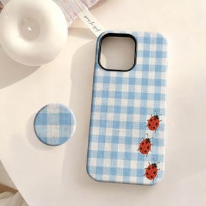 May include: A light blue and white gingham patterned phone case with three red ladybug illustrations. A matching pop socket is next to the phone case. A white candle is in the background.
