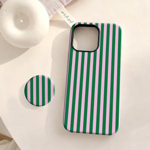 Puede incluir: Una funda de teléfono a rayas verdes y rosas y un agarre para teléfono a juego. La funda tiene un borde negro para la lente de la cámara. El agarre del teléfono es un disco redondo con el mismo patrón de rayas verdes y rosas.