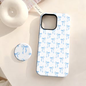 May include: A white phone case and matching pop socket featuring a repeating pattern of light blue bows and small white dots. The phone case has a black border around the camera cutout.