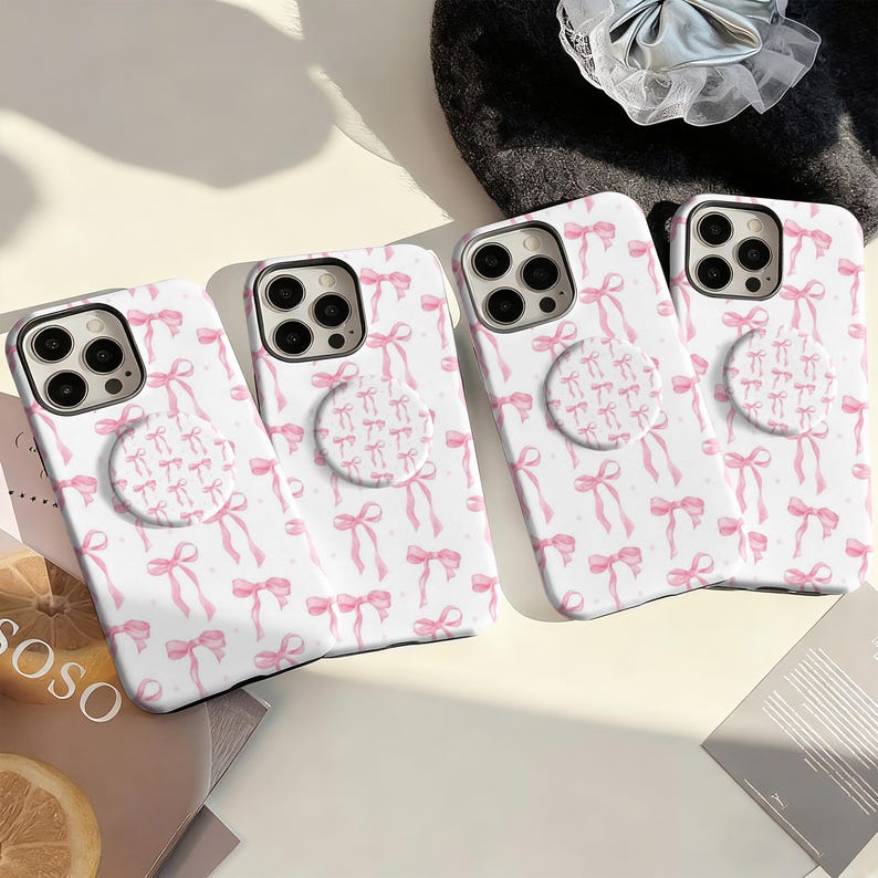 May include: Four white phone cases with a pink bow pattern and matching pop sockets. The cases have a repeating design of pink bows and small dots. The pop sockets also feature a bow pattern. The cases are for a smartphone.