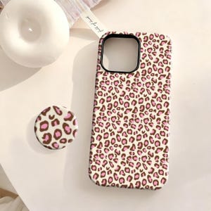 May include: A phone case and matching phone grip featuring a leopard print design in brown and pink on a cream background. The phone case has a black border around the camera cutout. The phone grip is round.