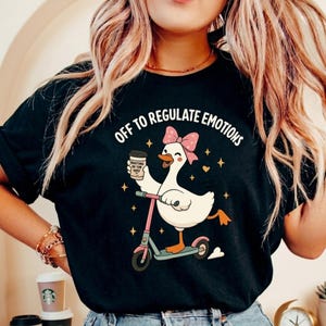 May include: Black t-shirt with the text "OFF TO REGULATE EMOTIONS". The graphic features a cartoon goose with a pink bow riding a pink scooter and holding a coffee cup. Gold stars and hearts are also in the design.