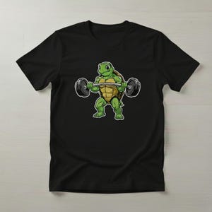Op de afbeelding: Zwart T-shirt met een cartoon schildpad die gewichten heft. De schildpad is groen en geel, met een vastberaden uitdrukking. De halter heeft zwarte gewichten aan elk uiteinde. Een leuk ontwerp voor fitnessliefhebbers.