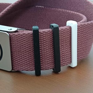 Peut inclure: Une ceinture en toile bordeaux avec une boucle argentée ornée d'un logo noir et blanc. La ceinture comporte deux passants noirs et un passant blanc. La ceinture est en toile résistante.