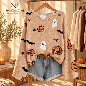 May include: A beige long-sleeve knit jumper featuring a repeating pattern of orange pumpkins, white ghosts, black bats, and small black stars. The jumper is displayed on a wooden hanger above a pair of light blue denim shorts with a brown belt.