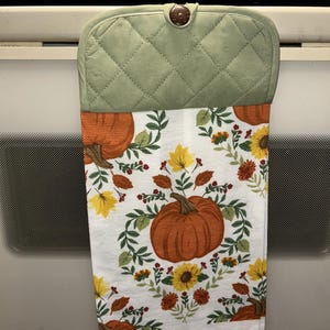 May include: A hanging kitchen towel featuring a white background with a pattern of orange pumpkins, yellow maple leaves, sunflowers, and green foliage. The top section is a sage green quilted fabric with a single dark brown button closure.