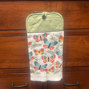 May include: A green quilted kitchen towel holder with a button closure. The attached towel is white with colorful butterflies and green leaf designs. The holder is hanging on a wooden dresser.