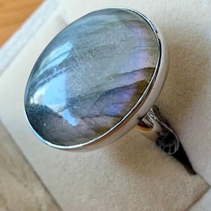Puede incluir: Un anillo de plata con una gran piedra de labradorita ovalada. La piedra muestra destellos iridiscentes de azul, morado y verde sobre una base gris. El anillo está engastado en una banda de plata pulida.