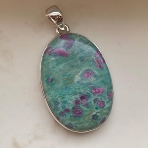 May include: Oval pendant featuring a green and pink gemstone set in a silver-colored metal frame. The gemstone has a mottled appearance with a mix of green and pink hues. The pendant has a small loop at the top for a chain.
