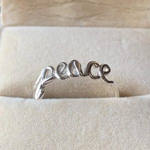 Puede incluir: Un anillo plateado con la palabra "peace" escrita en cursiva. El anillo se muestra en una caja de joyería beige, mostrando su acabado pulido y diseño elegante. La palabra "peace" es el punto focal.