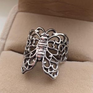 May include: A silver-colored ring featuring a detailed openwork design of a bee. The ring's body is a solid band, while the bee's wings and body are intricately cut out, creating a delicate and airy appearance. The ring is set against a neutral background.