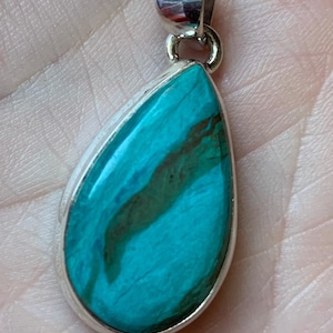 May include: A teardrop-shaped pendant with a vibrant turquoise stone and brown veining, set in a silver-colored metal frame. The pendant has a loop at the top for a chain.