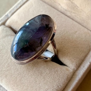 May include: A silver ring with a large, oval-shaped gemstone. The gemstone has a mix of dark purple, blue, and green colors. The ring is set in a silver band and is displayed in a beige ring box.