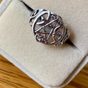 May include: A silver ring with an openwork design featuring a five-pointed star within a circular frame. The ring is set against a beige velvet background, showcasing its intricate details and craftsmanship.