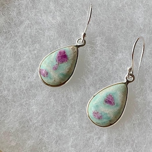 May include: A pair of teardrop earrings. Each earring has a pale turquoise stone with pink flecks and a silver metal border. The earrings hang from silver hooks, showcasing a unique and elegant design.