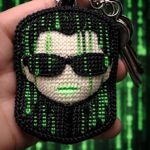 Puede incluir: Llavero de ganchillo hecho a mano con un rostro con gafas de sol, inspirado en la película Matrix. El llavero es negro, blanco y verde, con un llavero plateado y varias llaves adjuntas. El diseño es una representación estilizada de un personaje.