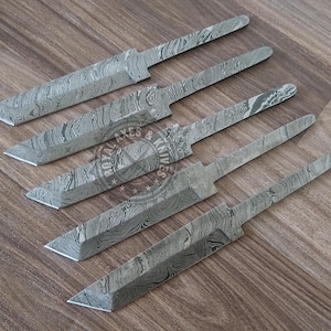 Könnte beinhalten: Fünf Damaststahl-Messerklingen mit einem einzigartigen Muster, angeordnet auf einer Holzoberfläche. Die Klingen haben ein silbernes und graues Farbschema, mit einem sichtbaren Logo in der Mitte. Die Messerklingen sind bereit für die Griffbefestigung.