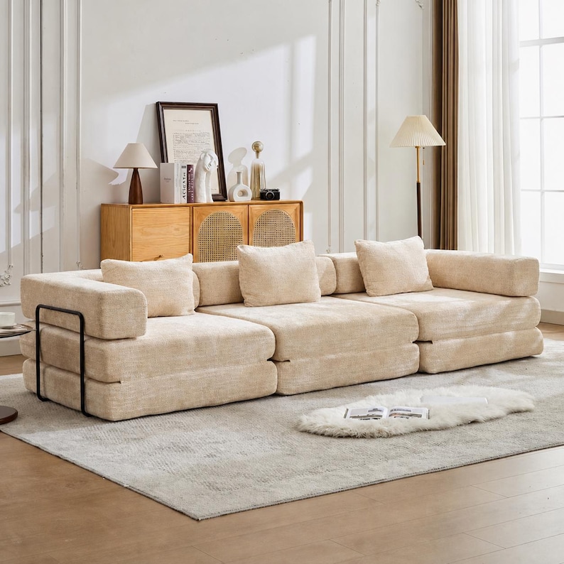 May include: A modular beige sofa with a textured fabric finish, featuring three low-profile seat sections, matching square back cushions, and a black metal frame accent on the left side, positioned on a light-coloured area rug in a bright room.