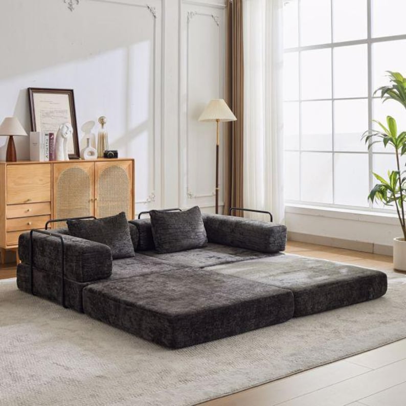 May include: A low-profile modular sofa bed upholstered in dark grey textured fabric. The piece features a black metal frame, two square back cushions, and a wide, flat seating area that extends forward to form a large, rectangular sleeping surface.