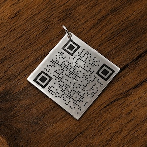 May include: A square silver-toned metal pendant featuring a black laser-engraved QR code pattern on its front surface. The pendant includes a small circular metal jump ring attached to the top corner for hanging, displaying a clean, flat design.