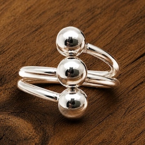 May include: A polished silver ring featuring a bypass design with three smooth, spherical metal balls arranged in a vertical line. The band is composed of two separate, curved silver wires that overlap, creating an open, adjustable structure.