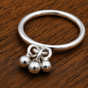 Puede incluir: Un anillo de plata pulida con una banda circular fina y lisa. En la parte inferior de la banda hay tres pequeños dijes esféricos de plata, cada uno suspendido de una diminuta argolla, creando un aspecto colgante similar a una campana.