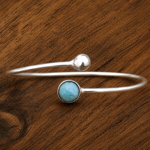 May include: A silver bypass bracelet featuring a polished round silver bead on one end and a round, light blue, marbled stone cabochon on the other. The open-ended design is crafted from a smooth, reflective silver band that curves into a circular shape.