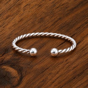 Può includere: Un bracciale a fascia color argento caratterizzato da una texture intrecciata simile a una corda lungo tutto il cerchio. Il bracciale è aperto e termina su entrambi i lati con una sfera liscia e lucida in metallo.